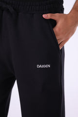 Black sweatpants with a white embroidered 'DAIGEN' logo on the left thigh, shown on a model with a hand in the pocket against a white background.
