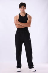 A man modeling black sweatpants and a black tank top, standing with arms crossed against a light gray studio background.