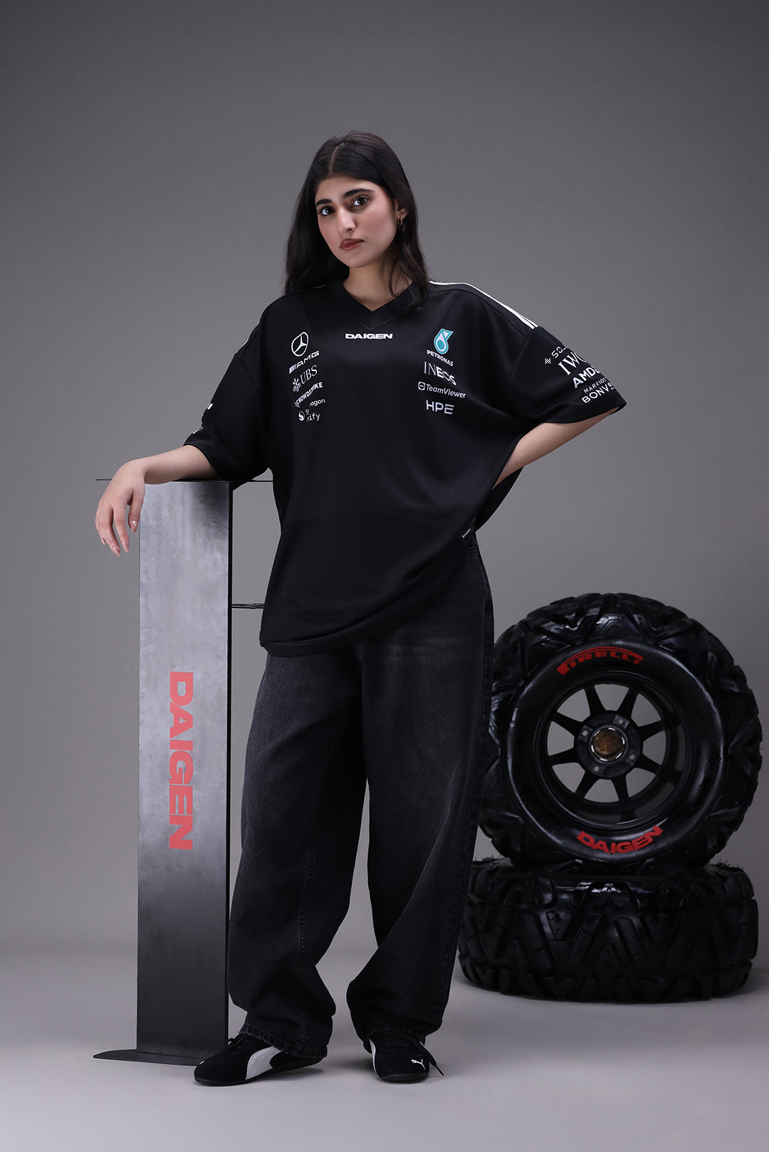 Person wearing a black oversized jersey with sponsor logos, standing next to a skateboard and formula 1 pirelli tires on a gray background